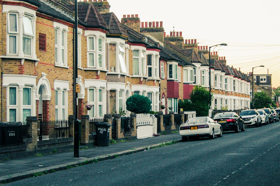 A row of traditional terraced houses on Goldhawk Road with front gardens and paved pathways, featuring neatly trimmed bushes and small trees. The houses have bay windows with white framing, some with